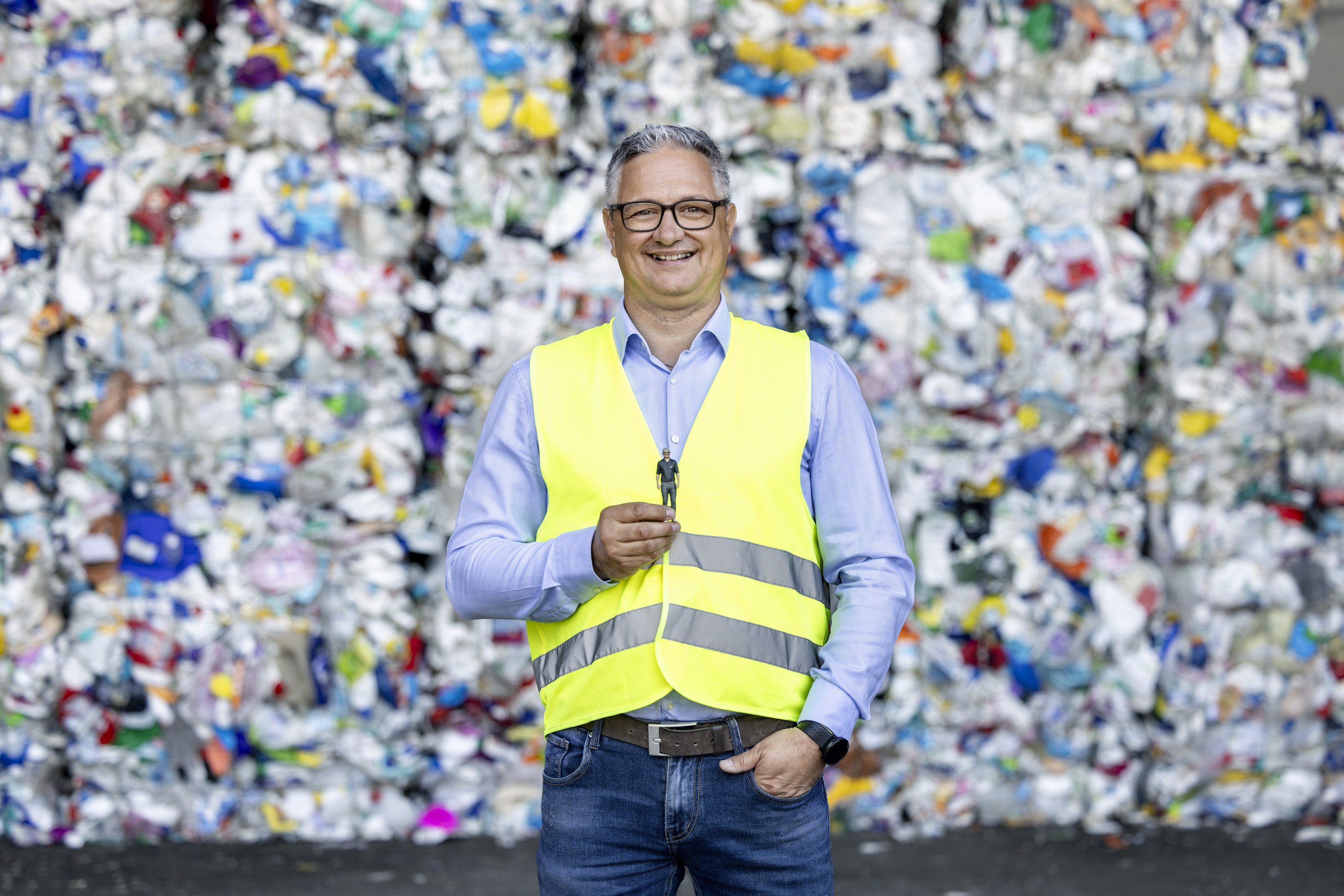 Ein Mann mit Brille, blauem Hemd und gelber Warnweste steht lächelnd vor einer Wand aus Recyclingballen. Er hält eine winzige Miniaturfigur zwischen Daumen und Zeigefinger vor seine Brust. Seine linke Hand steckt locker in der Tasche seiner Jeans.