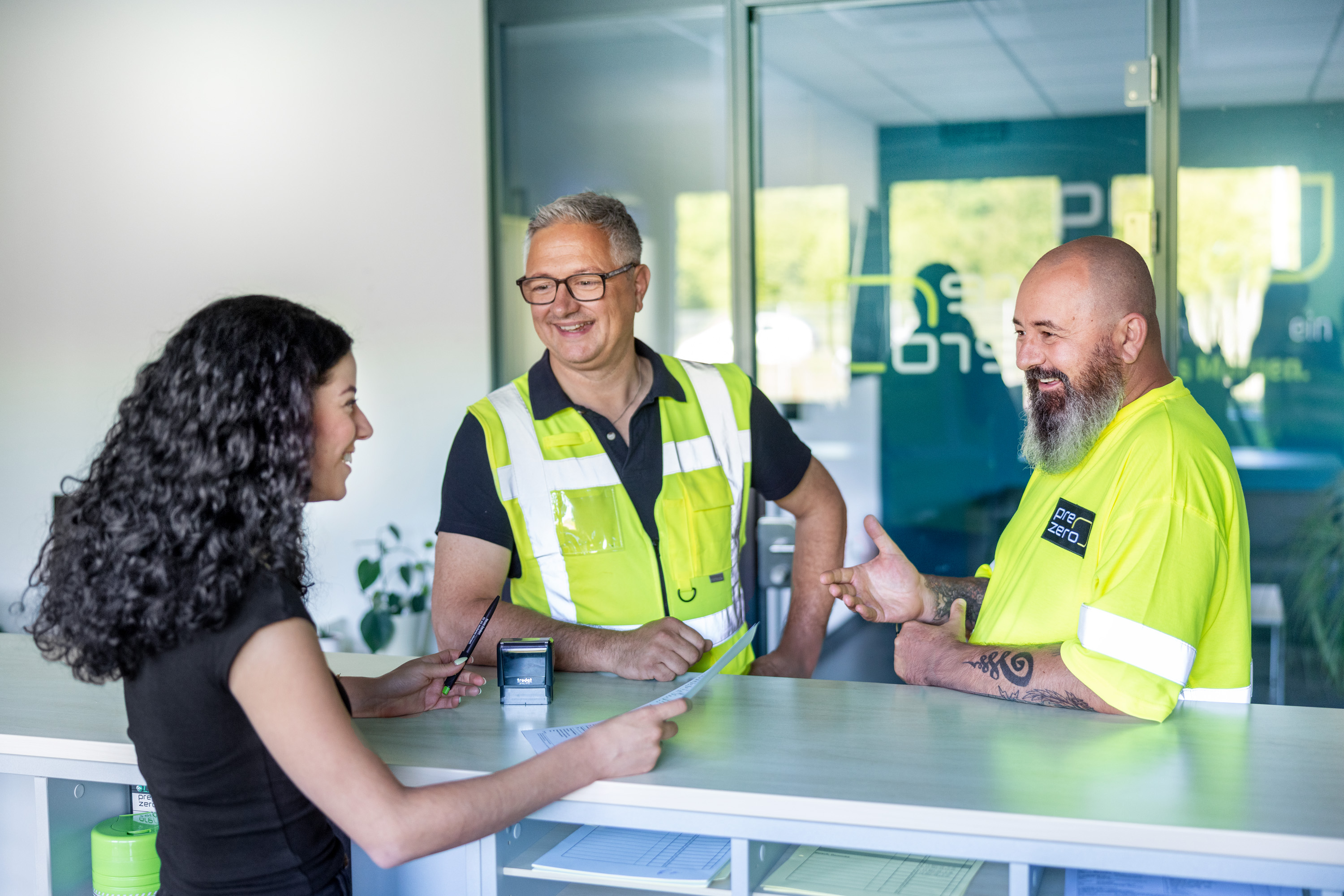 Drei Personen stehen lächelnd an einem Tresen in einem Büro- oder Empfangsbereich und unterhalten sich. Eine Frau mit dunklen, lockigen Haaren steht im Profil links hinter dem Tresen und hält einen Stift sowie ein Dokument in der Hand. Ihr gegenüber stehen zwei Männer in gelber Warnkleidung; einer trägt eine Brille und eine Weste über einem dunklen Poloshirt, der andere ein T-Shirt und hat einen markanten Bart sowie Tätowierungen am Arm. Die Atmosphäre wirkt freundlich und kollegial.