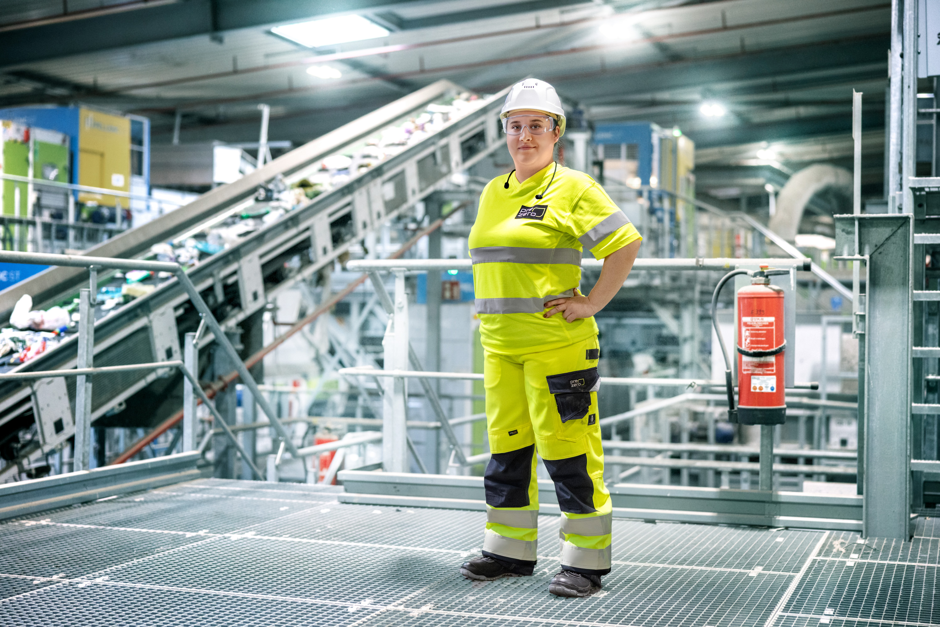 Eine Frau in neongelber Arbeitskleidung, weißem Schutzhelm und Schutzbrille steht mit in die Hüfte gestemmter Hand auf einem Gitterpodest in einer großen Industriehalle. Hinter ihr verläuft ein langes, schräg ansteigendes Förderband, das Wertstoffe transportiert. Ein Feuerlöscher ist rechts am Geländer montiert.