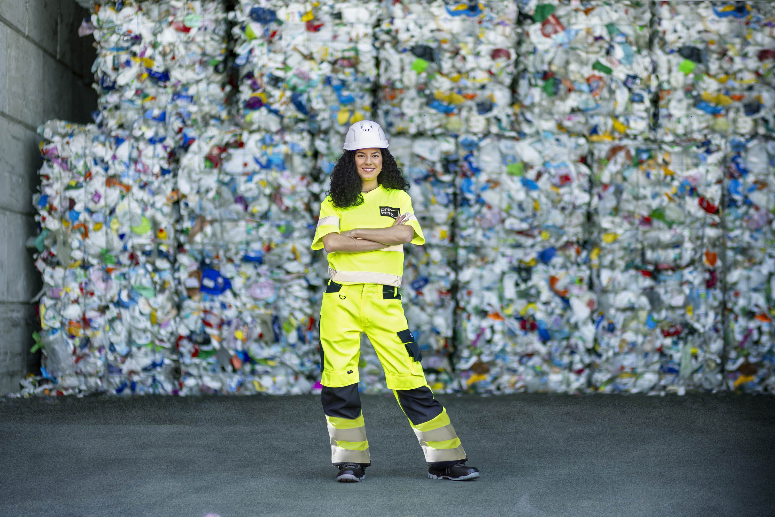 Eine Frau mit dunklen Locken steht selbstbewusst mit verschränkten Armen vor einer hohen Wand aus gestapelten Kunststoff-Recyclingballen. Sie trägt eine vollständige PSA (Persönliche Schutzausrüstung), bestehend aus einem weißen Schutzhelm sowie einer neongelben Arbeitshose und einem T-Shirt mit Reflexstreifen.