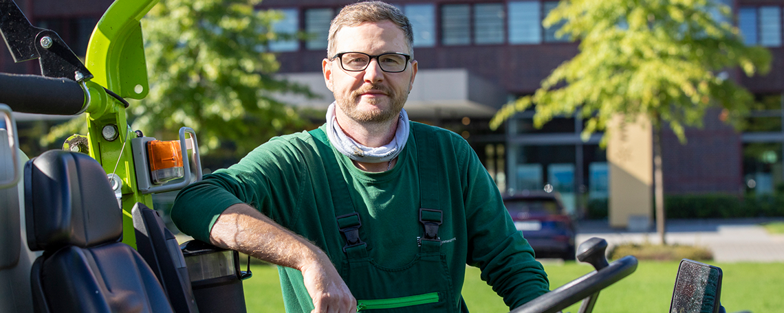 Zwei Männer gehen nebeneinander auf einem gepflasterten Weg in einem VIVAWEST-Quartier. Beide tragen grüne Arbeitskleidung und Schutzausrüstung: Schutzhelme mit Visier und Gehörschutz sowie Arbeitshandschuhe. Der Mann links trägt eine grüne Weste und hält einen Rechen in seiner rechten Hand. Der Mann rechts trägt ein grünes T-Shirt und hält eine Motorsäge in seiner linken Hand. Im Hintergrund sind Wohngebäude, Hecken und Bäume zu sehen.