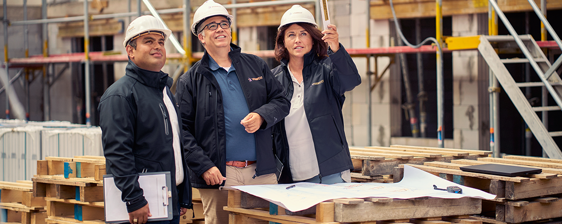 Das Bild zeigt eine Szene auf einer aktiven Baustelle. Drei Personen stehen eng beieinander, alle tragen weiße Schutzhelme und dunkle Jacken, was auf die Einhaltung von Sicherheitsvorschriften hinweist. Vor ihnen liegt auf einem improvisierten Tisch aus Holzpaletten ein großer Bauplan, der ausgebreitet ist. Die linke Person hält zusätzlich ein Klemmbrett in der Hand und die rechte Person zeigt mit einem Zollstock auf ein Objekt, was man auf dem Bild jedoch nicht erkennt. Es sieht so aus, als würde sie etwas erklären oder Anweisungen geben. Die dritte Person in der Mitte hört aufmerksam zu und richtet den Blick ebenfalls auf das Objekt. Im Hintergrund sind Gerüste, Baumaterialien und Teile eines bereits begonnenen Gebäudes zu erkennen, was die Atmosphäre einer laufenden Bauphase vermittelt. Die Szene wirkt konzentriert und koordiniert: Es ist ein Moment der Zusammenarbeit, in dem Fachleute gemeinsam die nächsten Schritte besprechen. Die gesamte Stimmung des Bildes vermittelt Organisation, Teamarbeit und den praktischen Alltag auf einer Baustelle.