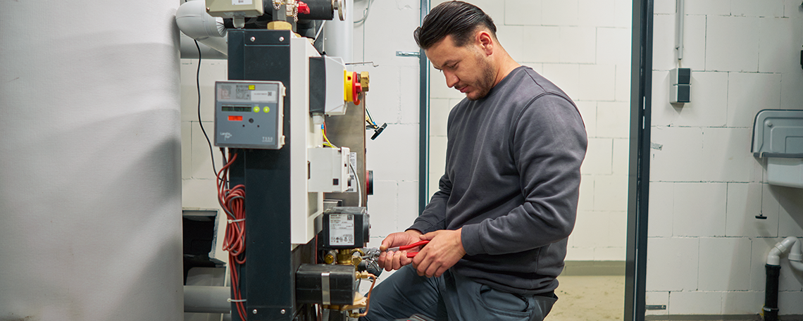 Ein junger Mann mit kurzem, dunklem Haar arbeitet konzentriert an einer technischen Anlage. Er trägt ein hellgraues Poloshirt und weiße Arbeitshandschuhe. Mit beiden Händen hält er ein größeres, professionelles Werkzeug, mit dem er eine Verbindung an einem Rohrsystem bearbeitet. Die Rohre sind teils aus Kupfer, teils mit Isolierungen versehen, und gehören zu einer Heizungs- oder Sanitärinstallation. Im Hintergrund ist eine unverputzte weiße Ziegelwand zu sehen.