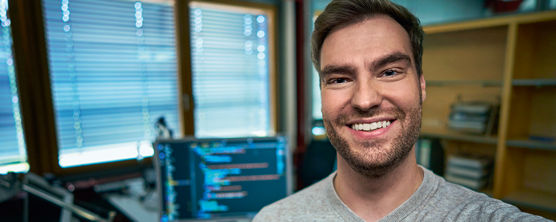 In dem Bild ist ein Mann zu sehen, der freundlich lächelt und in einem modernen Raum steht. Die Atmosphäre wirkt ruhig und konzentriert. Hinter der Person steht ein Computerbildschirm, auf dem ein Code-Editor geöffnet ist – deutlich erkennbar durch die farblich hervorgehobenen Programmierzeilen. Dies lässt darauf schließen, dass die Person gerade mit Softwareentwicklung oder Programmierung beschäftigt ist. Der Raum ist hell und wird von natürlichem Licht durch große Fenster durchflutet, deren Jalousien teilweise geschlossen sind. Rechts im Bild befindet sich ein Regal, in dem mehrere Gegenstände ordentlich gestapelt sind – vermutlich Bücher, Ordner oder andere Arbeitsmaterialien. Die Einrichtung wirkt funktional und aufgeräumt. Insgesamt vermittelt das Bild eine produktive und angenehme Arbeitsumgebung, die typisch für einen modernen Arbeitsplatz im Bereich Technologie oder IT ist. Die Kombination aus natürlichem Licht, technischer Ausstattung und freundlicher Ausstrahlung der Person schafft eine positive und motivierende Stimmung.