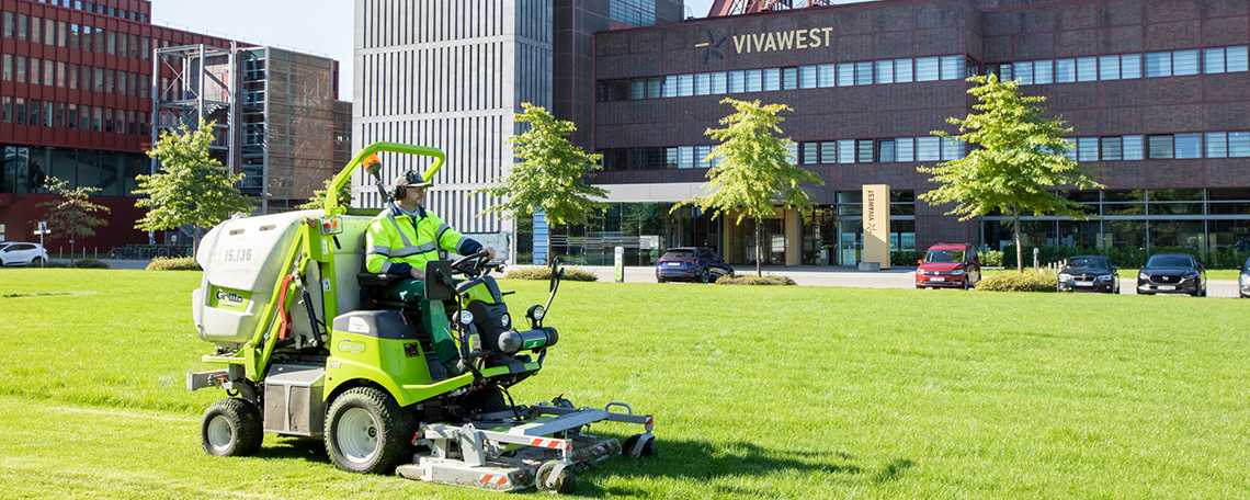 Zwei Männer gehen nebeneinander auf einem gepflasterten Weg in einem VIVAWEST-Quartier. Beide tragen grüne Arbeitskleidung und Schutzausrüstung: Schutzhelme mit Visier und Gehörschutz sowie Arbeitshandschuhe. Der Mann links trägt eine grüne Weste und hält einen Rechen in seiner rechten Hand. Der Mann rechts trägt ein grünes T-Shirt und hält eine Motorsäge in seiner linken Hand. Im Hintergrund sind Wohngebäude, Hecken und Bäume zu sehen.