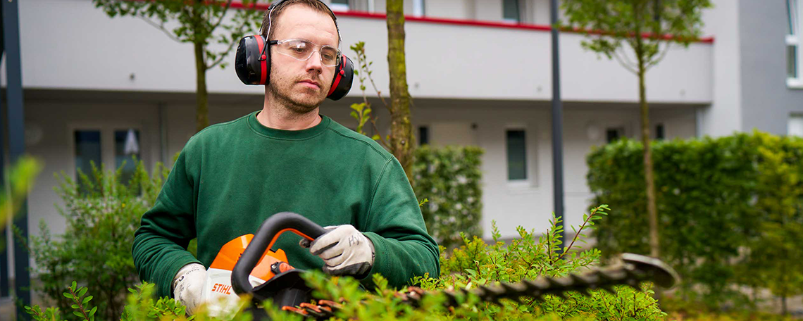 Ein junger Mann steht in der Mitte des Bildes und hält in beiden Händen eine Motorsäge von STIHL. Er trägt eine grüne Arbeitskleidung und Schutzausrüstung: Schutzbrille und Gehörschutz sowie Arbeitshandschuhe. Im Vordergrund ist eine Hecke zu sehen, die der Mann gerade schneidet. Im Hintergrund befindet sich ein Wohngebäude. Vor diesem Gebäude sind eine Hecke und drei Bäume zu sehen. 