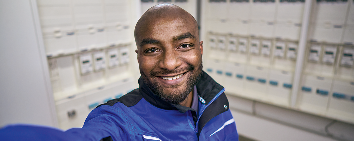 Ein junger Mann mit Glatze und einem Bart arbeitet lächelt in die Kamera. Er trägt eine blau-schwarze Arbeitsjacke. Im Hintergrund ist ein offener Elektroschaltschrank zu sehen.