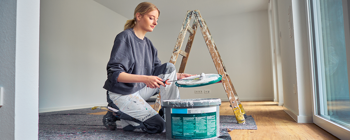 Das Bild zeigt eine junge Frau, die in der Mitte von einem Wohnraum kniet. Vor ihr steht ein Farbeimer mit weißer Farbe. In ihrer linken Hand hält sie den Deckel des Farbeimers und in der rechten Hand eine Farbrolle, mit der Sie über die Innenseite des Farbdeckels rollt. Im Hintergrund ist eine Stehleiter zu sehen. Der Fußboden ist zudem mit grauem Malerflies ausgelegt. Die Wände sind weiß gestrichen.