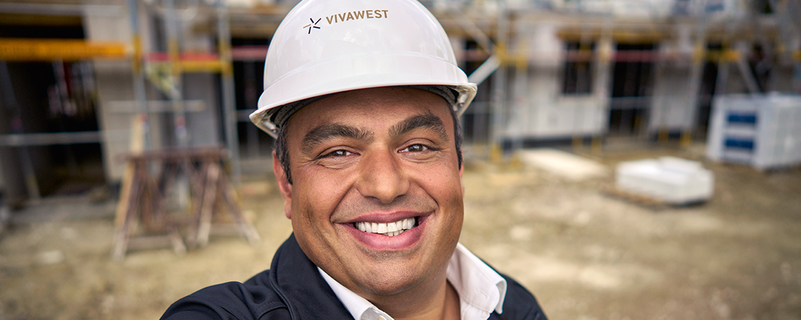 In dem Bild ist zentral ein Mann zu sehen, der freundlich lächelt und einen Bauhelm trägt. Der Helm ist weiß und hat das VIVAWEST Logo abgebildet. Im Hintergrund sind Gerüste, Baumaterialien und Teile eines bereits begonnenen Gebäudes zu erkennen, was die Atmosphäre einer laufenden Bauphase vermittelt. Die Kombination aus natürlichem Licht, technischer Ausstattung und freundlicher Ausstrahlung der Person schafft eine positive und motivierende Stimmung.