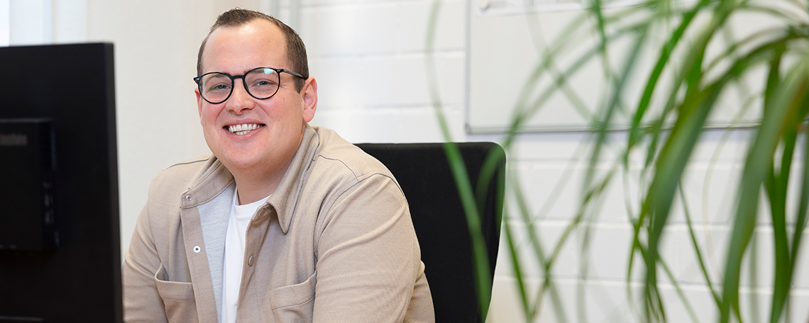 Ein Mann mit kurzen braunen Haaren und einer Brille steht an einem höhenverstellbaren Schreibtisch vor zwei großen Bildschirmen. Er trägt ein weißes Hemd und lächelt leicht, während er konzentriert auf die Bildschirme blickt. Im Hintergrund ist ein Fenster, durch das Tageslicht hereinfällt. Auf der Fensterbank steht eine grüne Pflanze.