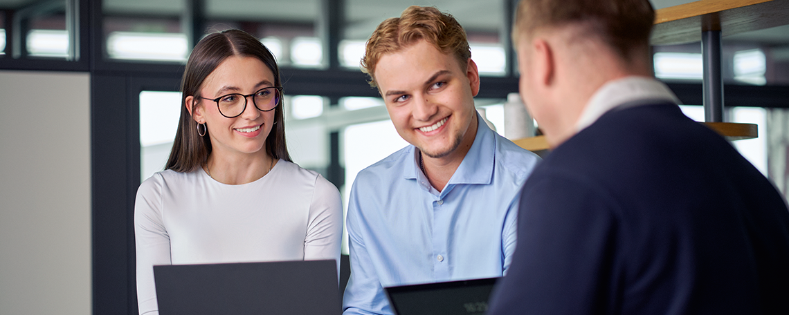 Das Bild zeigt eine junge Frau auf einer grauen Treppe mit einer grünen Bluse und langen dunklen Haaren. Sie lächelt freundlich in die Kamera und hält dabei einen Notizblock mit dem VIVAWEST - Logo in der Hand. Im Hintergrund stehen weitere Personen, jedoch sieht man nur die Arbeitskleidung und nicht die Gesichter. Links ist ein Mann mit einer blauen Jeans und einem schwarzen Jackett zu erkennen, sowie einen Elektroniker in blauer Arbeitskleidung. Rechts steht ein Gärtner mit grüner Arbeitskleidung und einem Tablet in der Hand. Hinter ihm kann man eine weitere Person mit orangefarbener Hose erkennen. 