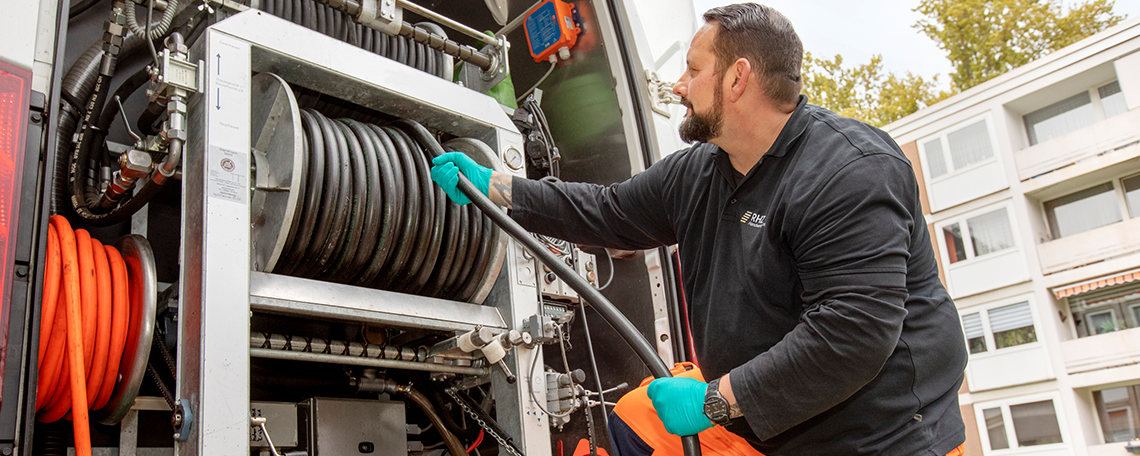 Das Bild zeigt einen jungen Mann mit orangefarbener Arbeitskleidung und grünen Handschuhen. Dieser lächelt freundlich in die Kamera. Er hockt auf gepflasterten Boden. Mit beiden seiner Hände hält er einen schwarzen Schlauch fest, welcher in Richtung Boden geht. Links neben ihm stehen zwei orangene Hütchen, wie eines vor ihm. Rechts neben ihm steht ein schwarzes Gerät mit einem lilafarbenen Kabel, welches eine Kanalinspektionskamera ist. Im Hintergrund steht ein weißer Transporter mit geöffneten Flügeltüren. Es befinden sich in dem Fahrzeug verschiedene Rohre und andere Werkzeuge.   