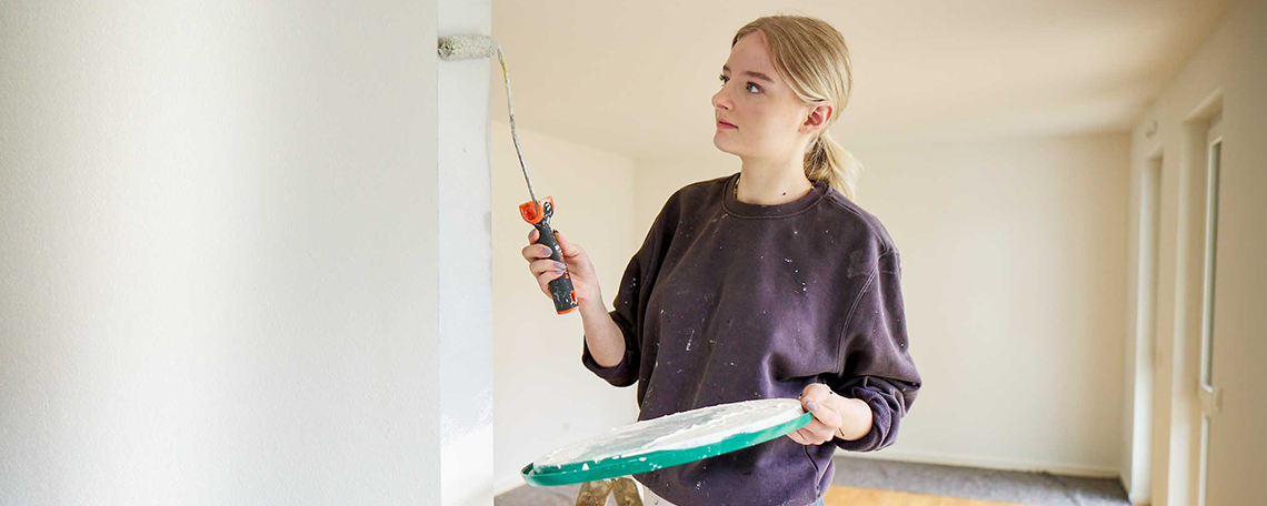 Eine in hellgrau gekleidete junge Frau hockt auf dem Boden. Neben ihr steht eine Leiter und vor ihr ein blauer Farbeimer, auf welchem eine Farbrolle liegt. 