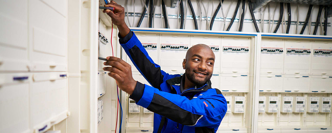Das Bild zeigt einen Mann, welcher ein hell graues Poloshirt trägt und hellbraune Haare hat. Er schaut auf einen offenen Zählerschrank in dem eine Leitung angeschlossen ist und hat dabei ein leichtes Lächeln auf den Lippen. In seinen Händen hält er einen zweipoligen Spannungsprüfer mit diesen er die anliegende Spannung misst. Im Hintergrund sieht man weiße Backsteinwände und einen weiteren weißen Zählerschrank sowie an der Decke eine Kabeltrasse.
