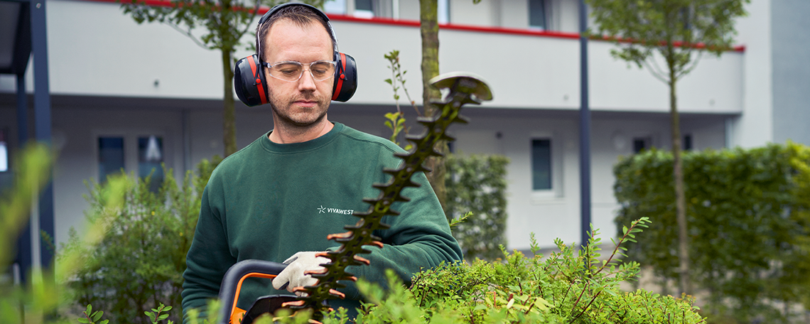 In der Mitte auf dem Bild ist ein junger Mann mit grüner Arbeitskleidung zusehen. Er kniet mirt einem Bein auf dem Boden und das andere hat er aufgestellt. In seiner linken Hand hält er eine rote Handsäge, mit welcher er einen langen Ast durchsägt. Deshalb schaut er nach unten auf den Ast. Im Hintergrund sieht man grüne Bäume und Büsche. 