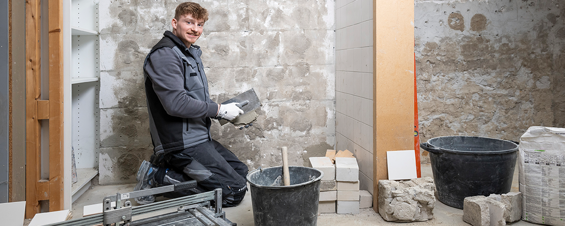 Das Bild zeigt eine kleine Nische mit grauen unbearbeiteten und ungefliesten Wänden. Im Vordergrund ist ein junger Mann in der Hocke mit einer schwarz-grauen Jacke und einer schwarzen Arbeitshose. Er lächelt freundlich in die Kamera. Vor sich in der Hand hält er eine weiße Fliese, welche er mit einem Glätter bestreicht. Hinter ihm befindet sich eine ungeflieste Wand. 