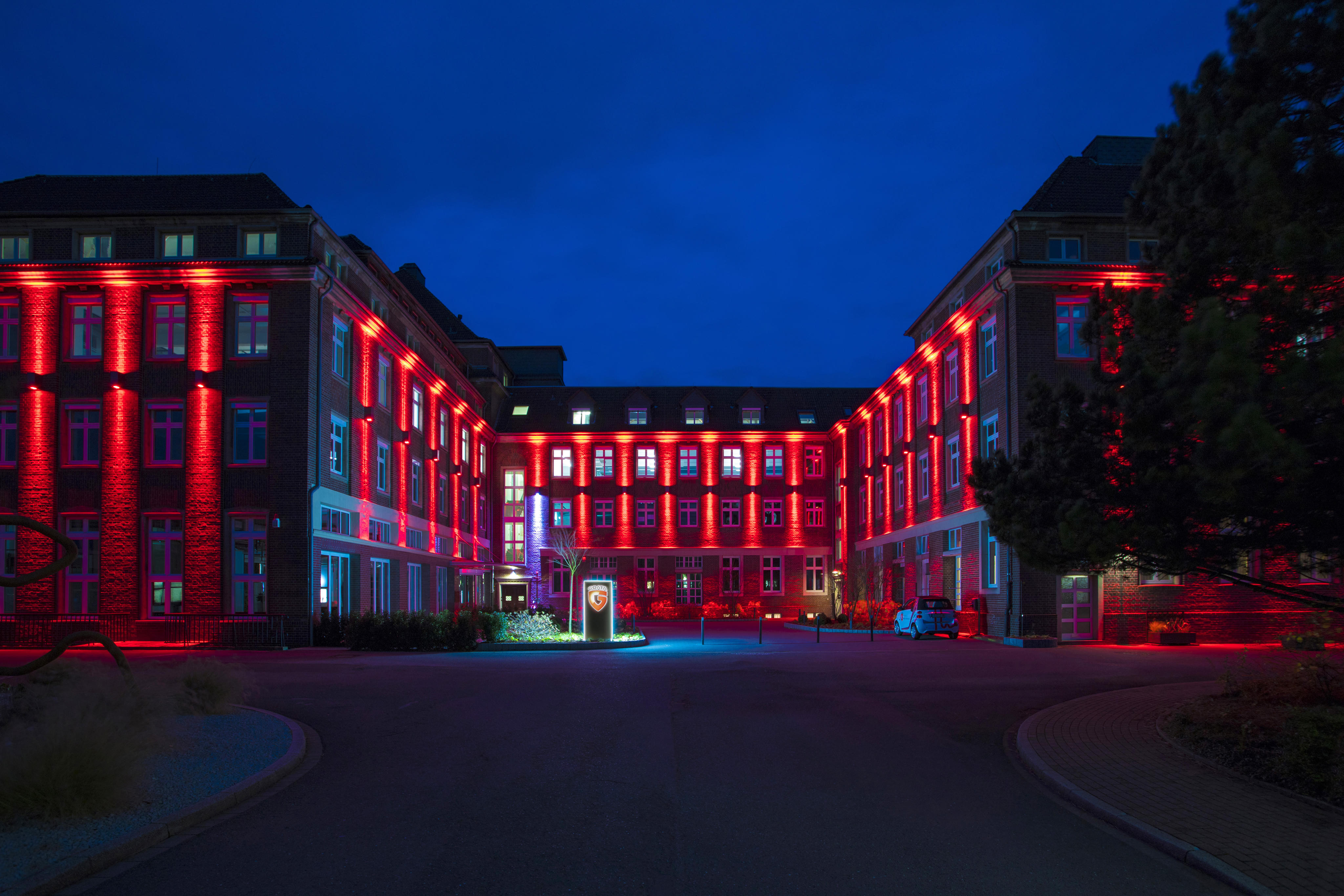 Das Bild zeigt den G DATA Campus bei Nacht: U-förmiges Gebäude in rotem Licht, mit Hofeingang in der Mitte, umgeben von dunklem Himmel und Bäumen.